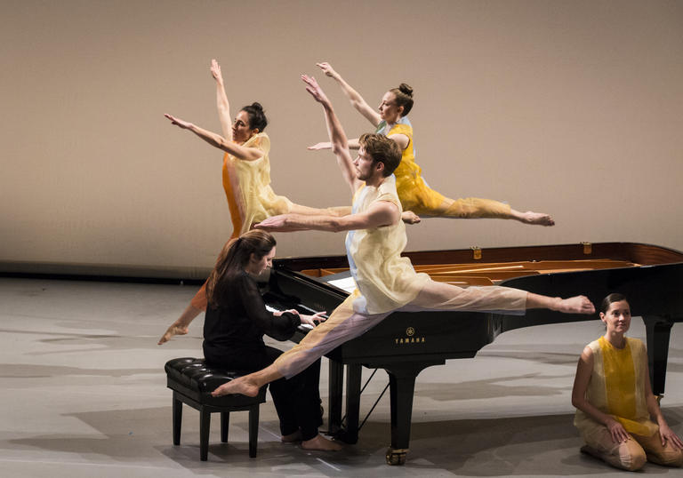 three dancers in yellow leap around a grand piano, one dancer is sat on the floor