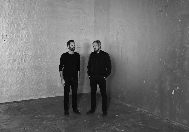 Dustin O'Halloran and Adam Wiltzie standing in a bare room, wearing all black.