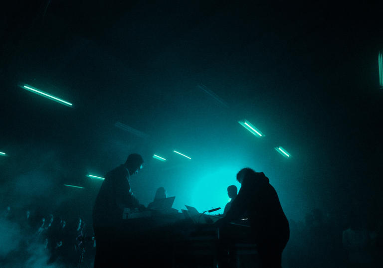 A live performance featuring four musicians facing each other with smoke and stobe lights around them