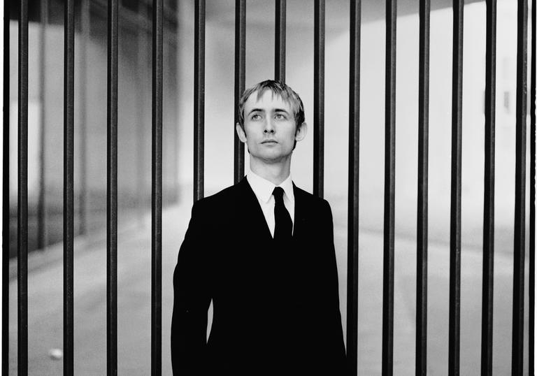 black and white photo of Neil Hannon wearing a suit against a backdrop of vertical bars