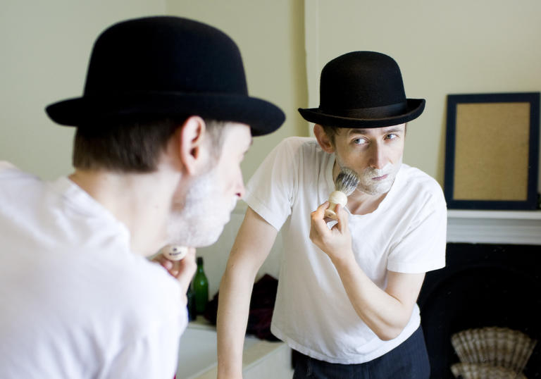Colour photo of Neil Hannon wearing a bowler hat, shaving with foam on his face