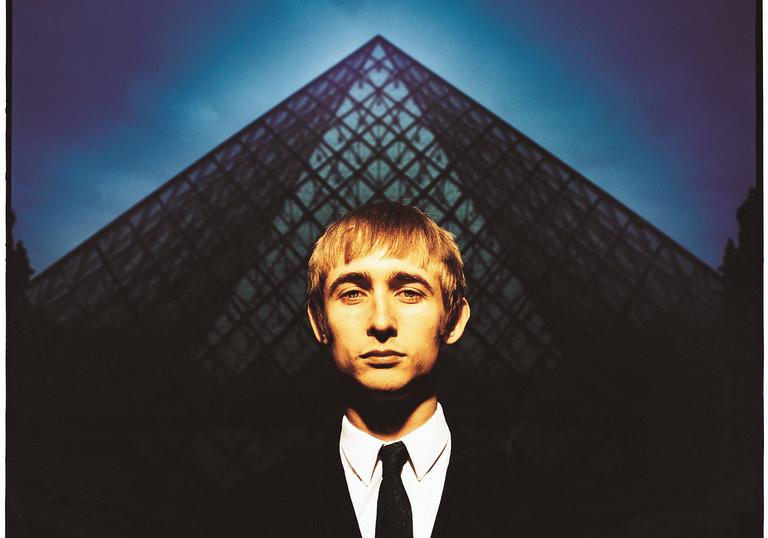 Neil Hannon in a black and white suit and tie standing in front of a blue pyramid