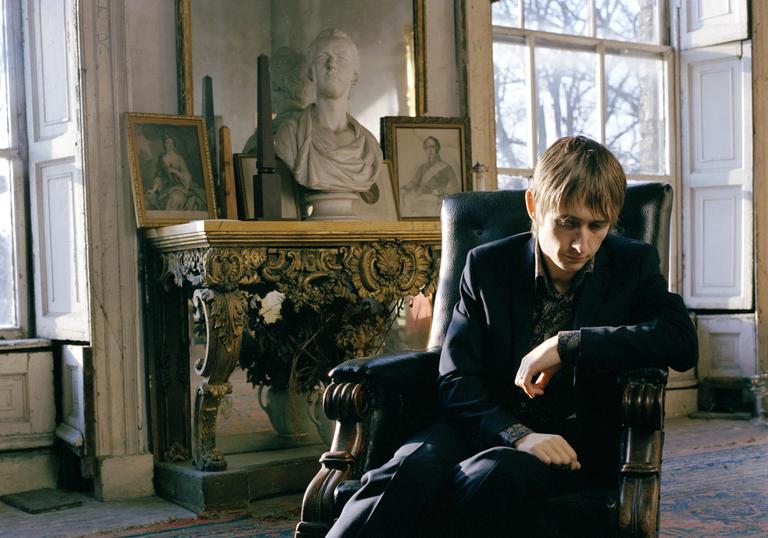 A colour photo of Neil Hannon lounging in a chair in a slightly dilapidated stately home, with antique furniture in the background