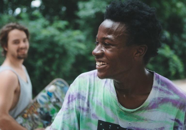Two men are laughing, one is in the background holding a skateboard