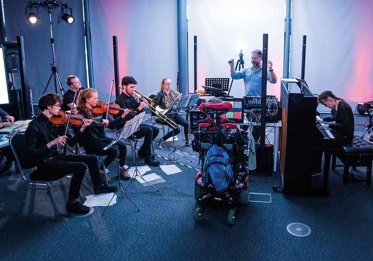 Group of young people disabled and non disabled are playing musical instruments and facing the conductor with their backs to the camera