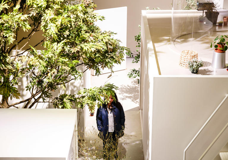 Woman walks through a gallery space with a tree and white houses