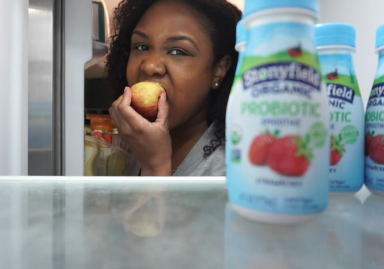a woman eating an apple looks into her fridge and sees some probiotic yogurts