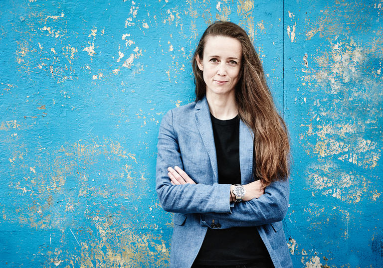 Conductor Jessica Cottis standing against a blue patterned wall with arms crossed