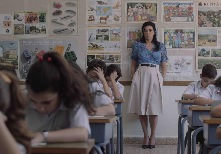 A teacher stands at the back of her classroom with children at desks