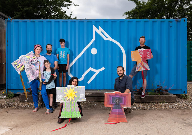 A group of people stand in front of a blue metal box with an image of a horse on it holding kites that they have made