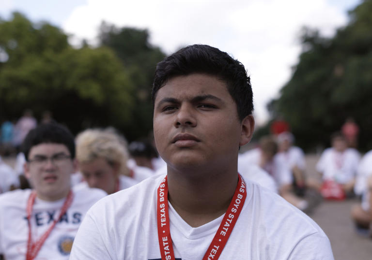 a boy in a white t shirt sits with many others behind him
