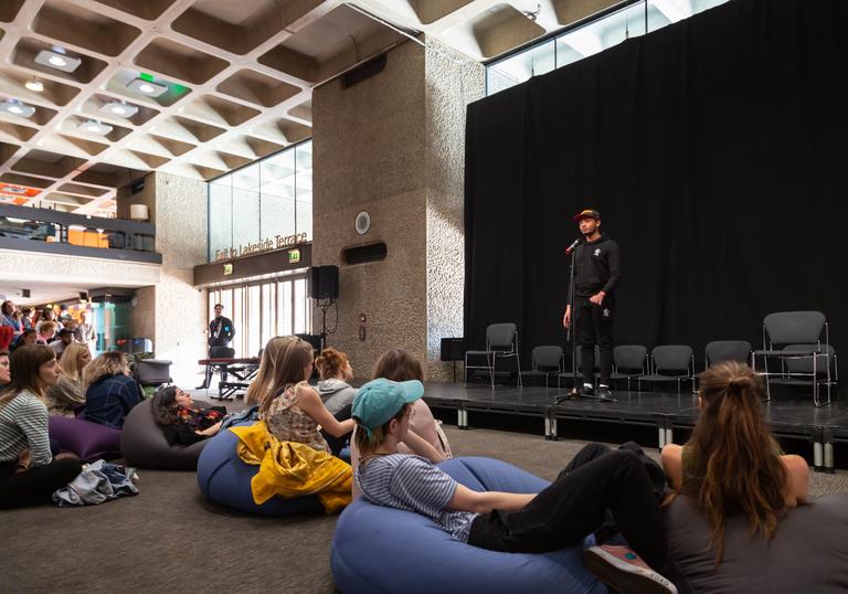 A group of people are sat on beanbags on the floor watching a person on stage