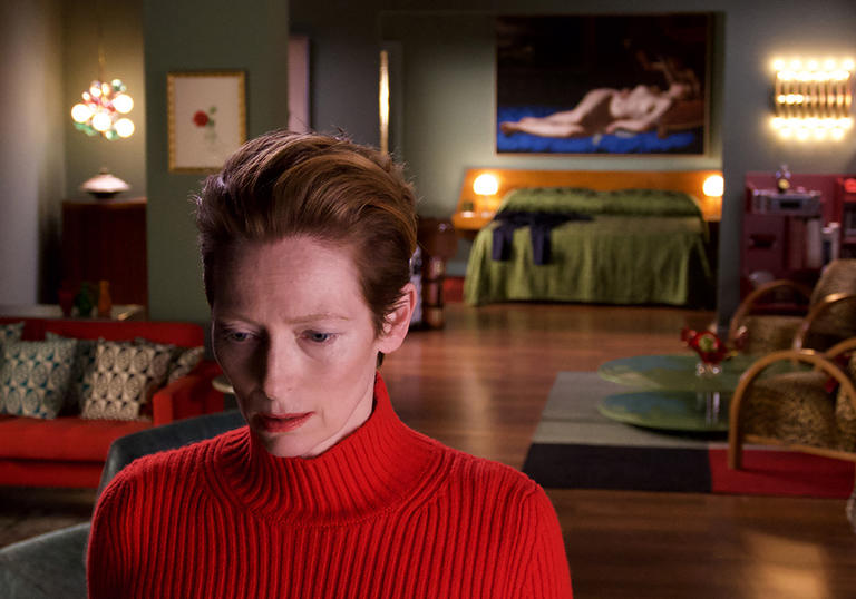 tilda swinton in a bright red top and swept back hair standing in an apartment