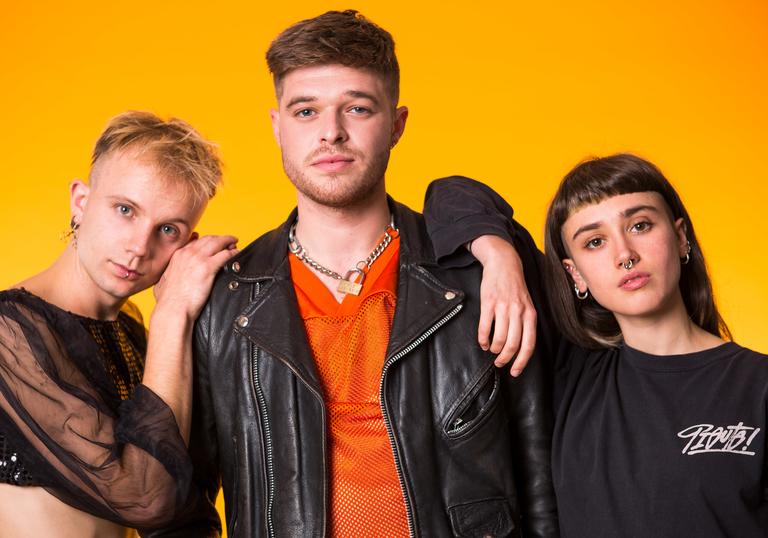 Three people stand together in front of an orange background wearing black clothes