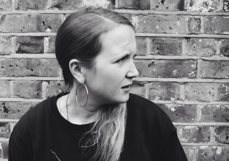 Hollie McNish leaning against a brick wall and looking over her left shoulder