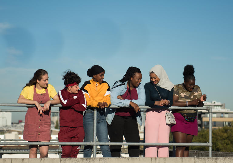 A group of six young girls stand in a row on what looks like the roof of a building. They are leaning on some railings and there's a clear blue sky above them.