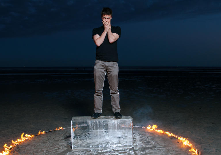 David Finnegan stands on an ice block, in a desert. A ring of fire burns around him.