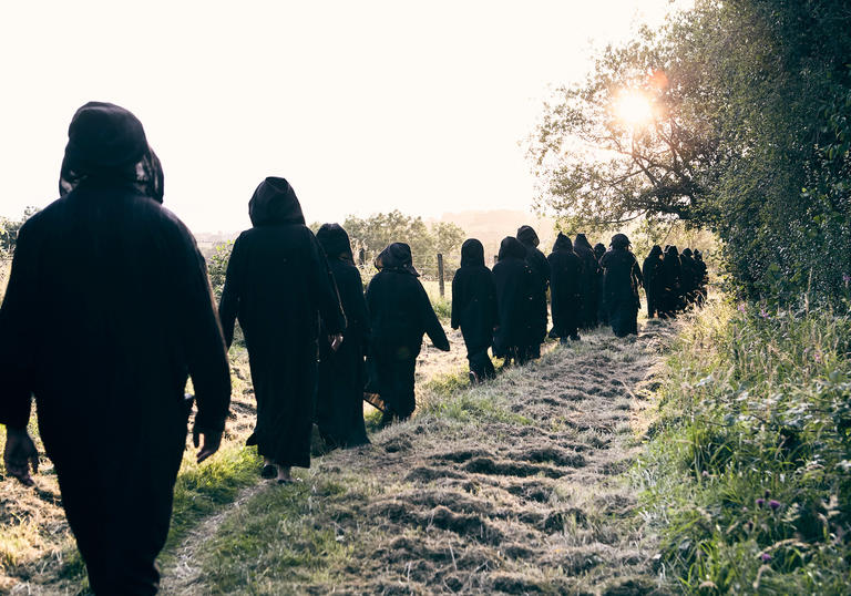 A row of people in black hooded long cloaks walk through a rural field in sunshine. We can't see their faces from behind.
