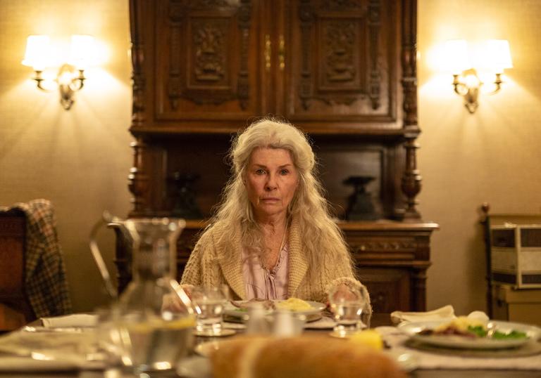 an old woman sits at the head of a long table with her long white hair cascading down