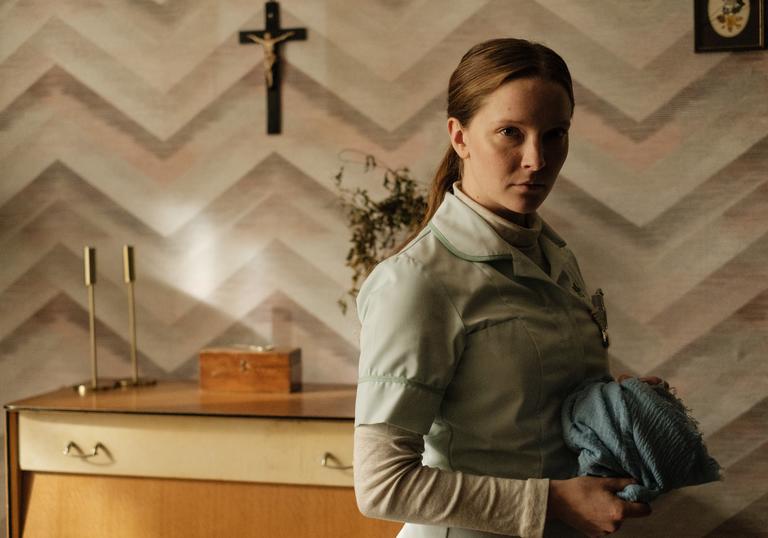 A woman stands looking toward the camera in front of a cross