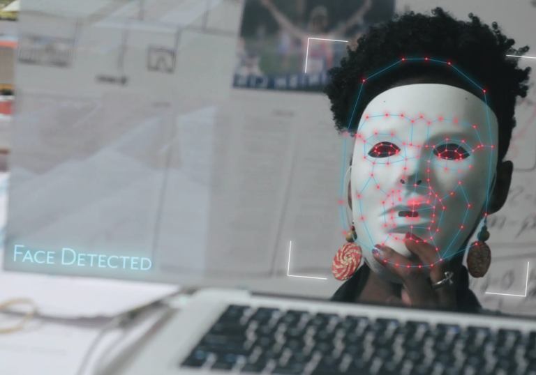 In front of a computer, a black woman holds up a white AI mask being detected by a computer, in Coded Bias