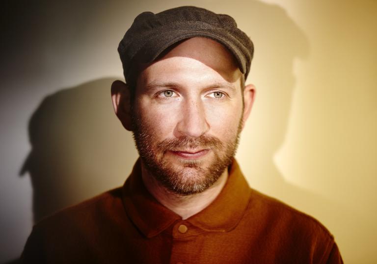 photo of Matthew Halsall wearing a cap and shirt against a plain backgound, being lit from multiple sources to create shadows