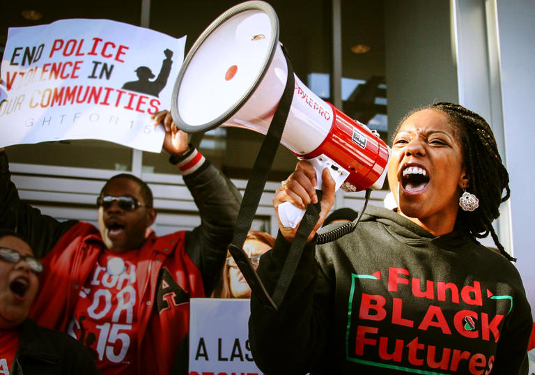 activists wearing BLM jumpers hold up signs and shout into a megaphone