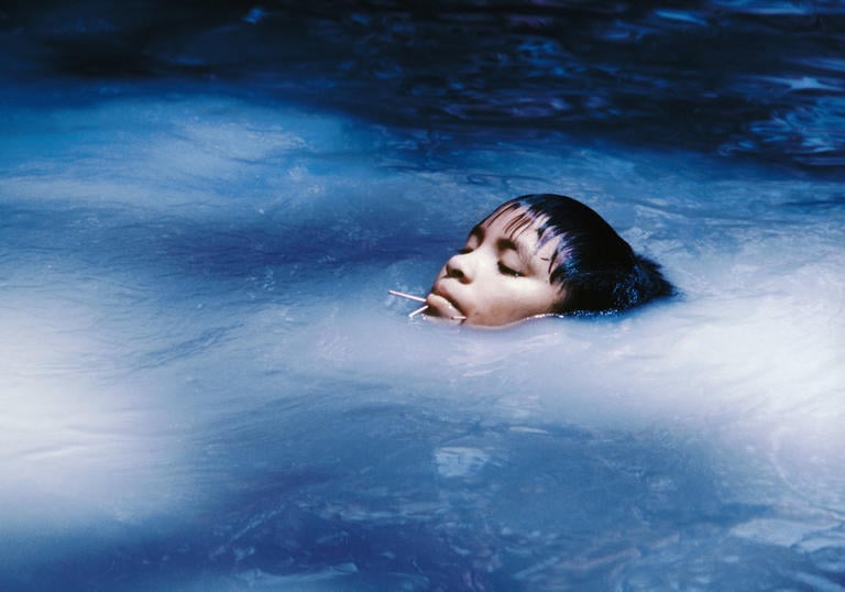 A young boy swims in blue water, his head partially emerging out of the surface