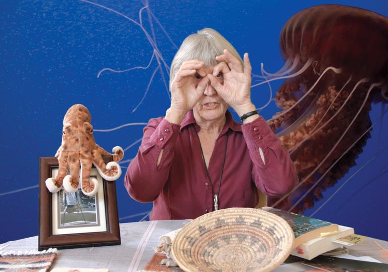 A photo of Donna Haraway sitting at a table. In the background a jelly fish floats in water