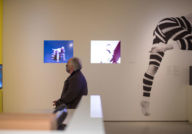 A man sitting on a white bench watching a television screen, behind him is a large photo of a dancer in a black and white patterned leotard