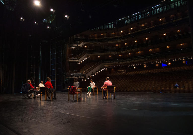 A wide shot of the Barbican stage with empty auditorium and a group of people sitting on the stage