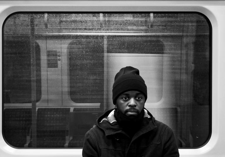black and white photo of Alpha Mist sitting in a tube carriage, he is wearing a big coat and a beanie hat, with a neutral expression