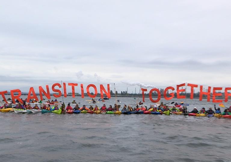 climate activists tie themselves together in kayaks holding a sign that says 'transition together'