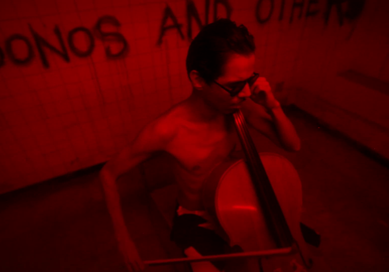 a man plays a cello in a dark room full of red light and graffiti on the wall