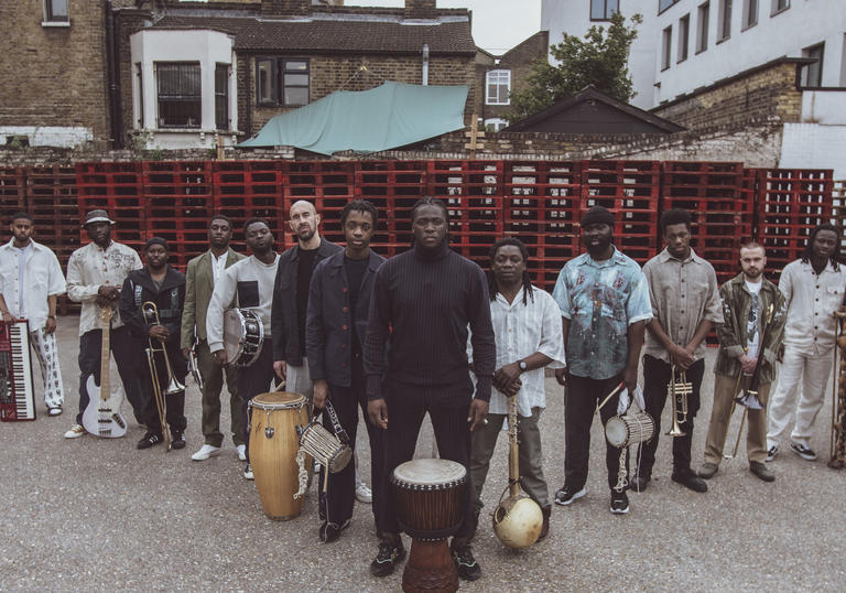 Balimaya Project standing in a triange formation with their instruments at their feet, in front of stacked red crates.