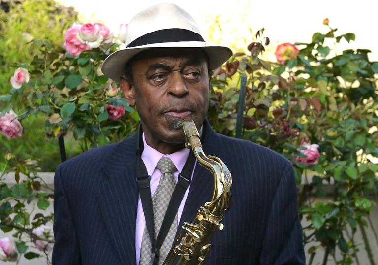Archie Shepp playing a saxophone. He is wearing a black suit with a white hat and standing in front of a rose bush.