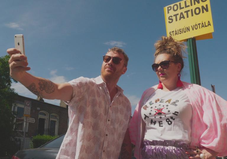 a man and a woman dressed in pink take a selfie with the polling station sign behind them