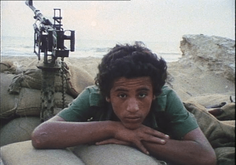 A young man lies on sand bags with a automatic gun behind him