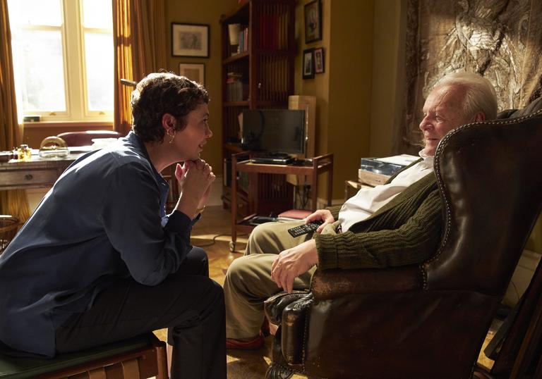 a daughter and her father sit looking at each other in a cluttered living room