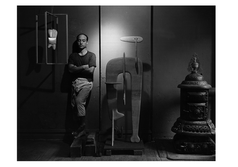 a black and white photo of a man standing against a wall, there are sculptures besides him