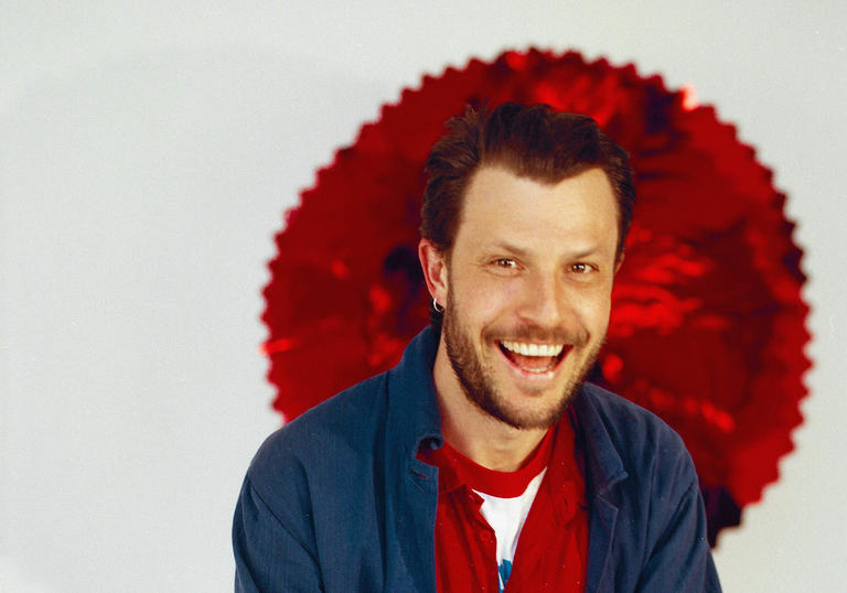 A smiling Orlando Weeks standing against a red and white background
