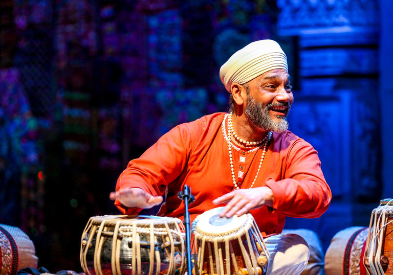 Sukhwinder Singh playing the tabla and smiling to his left. He is wearing an orange top, beaded necklaces and a white turban.