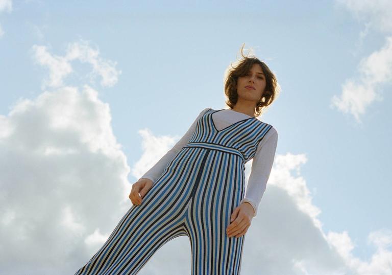 colour photo of Aldous Harding against a blue sky background. Photo is shot from below and she wears a blue striped jumpsuit