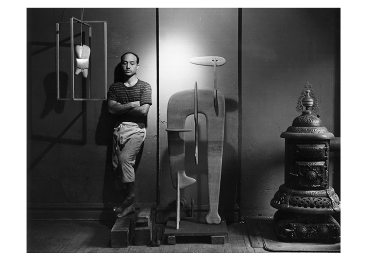Isamu Noguchi standing in his studio with arms folded