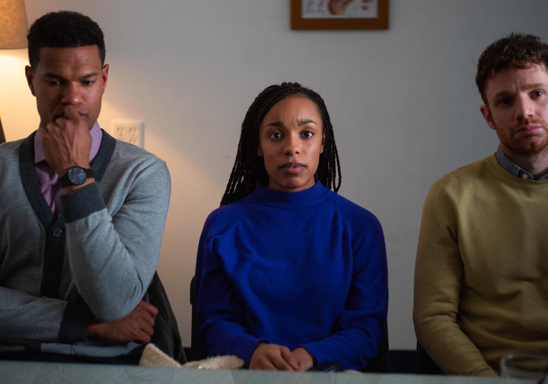 A woman sits between two men, all facing the same way, with thoughtful looks on their faces