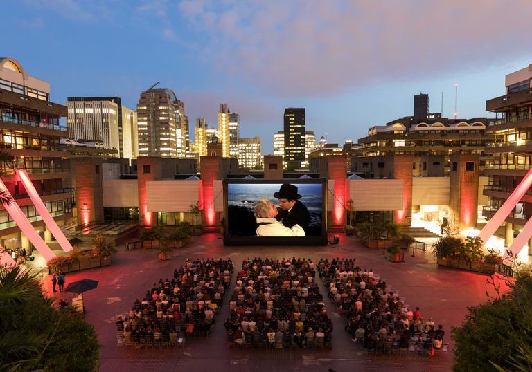 A wide shot from above of the Outdoor Cinema on the Sculpture Court of the Barbican Centre, with a seated audience facing a screen