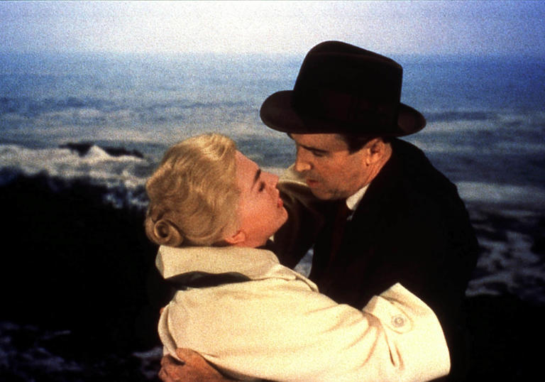 Kim Novak in a white coat and James Stewart in a black suit and hat embrace, in Alfred Hitchcock's Vertigo