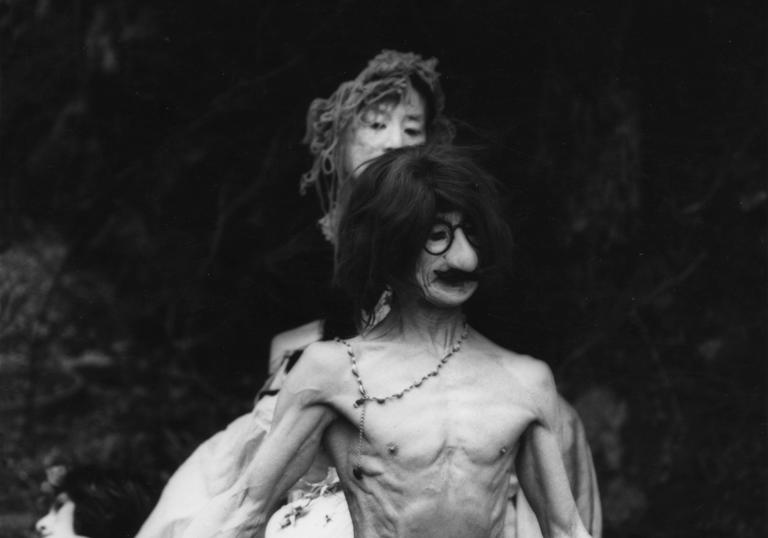 a man wearing an ugly mask stands topless with a woman behind him
