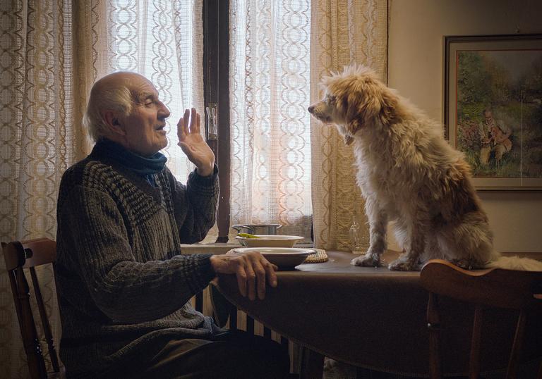 A old Italian man sits at a table and looks at his dog, who is sitting on the table
