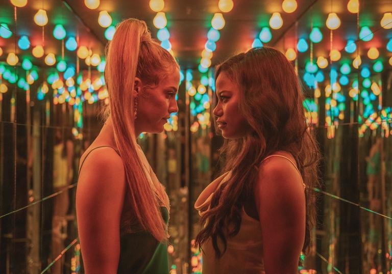 Riley Keough and Taylour Paige look directly at each other in a hallway of mirrors with multicoloured lights above